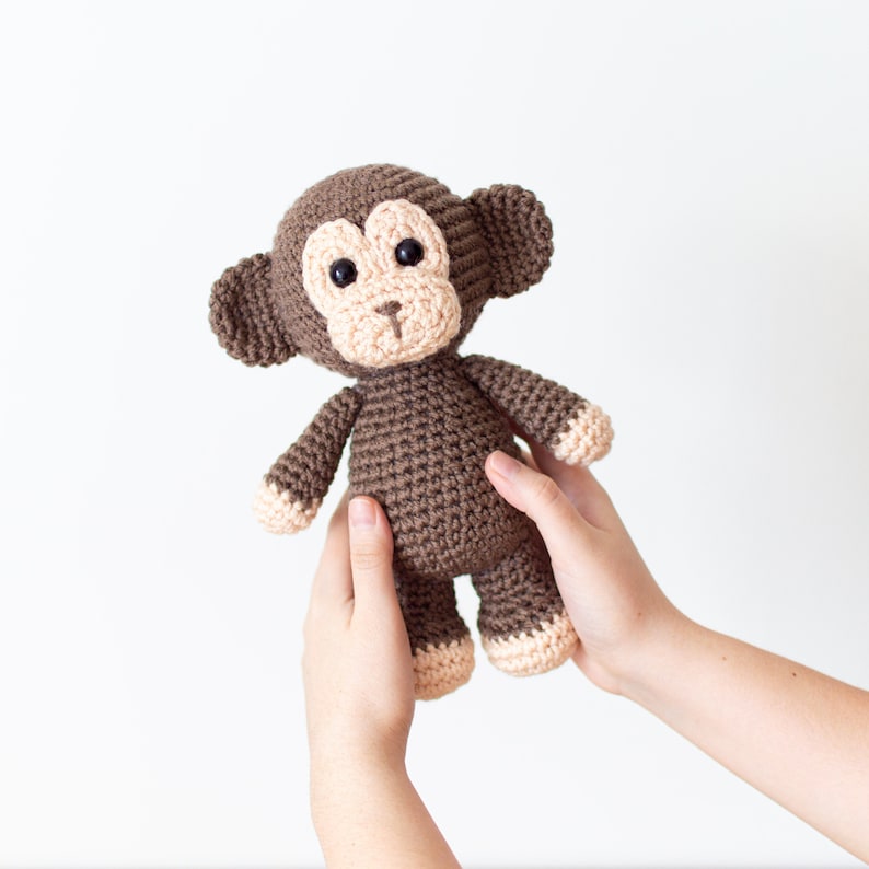 May include: A brown crocheted monkey toy with beige accents on the face and feet. The monkey is being held by two hands.