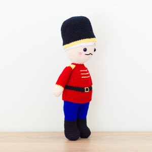 May include: A crocheted toy soldier with a black hat, red jacket, blue pants, and a yellow belt. The toy has a white face with black eyes, a black mustache, and a pink blush.