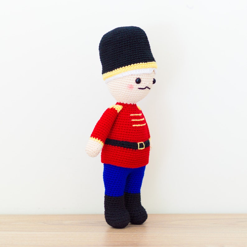 May include: A crocheted toy soldier with a black hat, red jacket, blue pants, and a yellow belt. The toy has a white face with black eyes, a black mustache, and a pink blush.