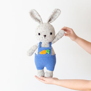 May include: A crocheted bunny toy wearing blue dungarees with a carrot patch. The bunny has white fur with brown speckles and black eyes.