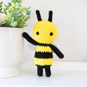 May include: A crocheted bee toy with black and yellow stripes, black legs, and white wings. The bee has a yellow head with black eyes and a pink blush.