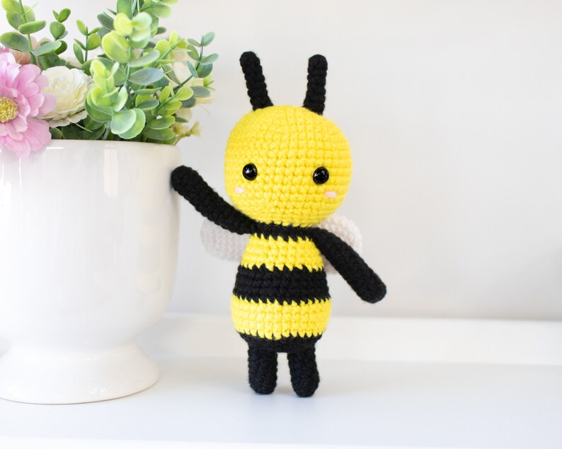 May include: A crocheted bee toy with black and yellow stripes, black legs, and white wings. The bee has a yellow head with black eyes and a pink blush.