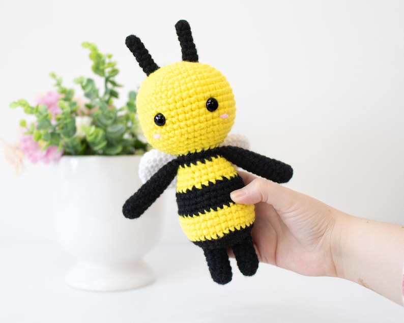 May include: A crocheted bee plush toy with black and yellow stripes, black antennae, and white wings. The bee has black eyes and a pink blush.