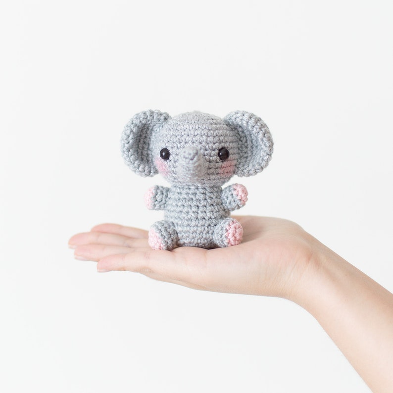 May include: A small grey crocheted elephant toy with pink accents on the ears and feet. The elephant is sitting in the palm of a hand.