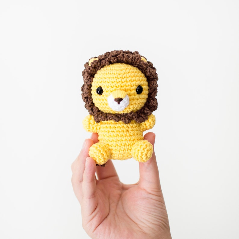 May include: A small crocheted lion toy with a yellow body and brown mane. The lion has black eyes and a white nose.