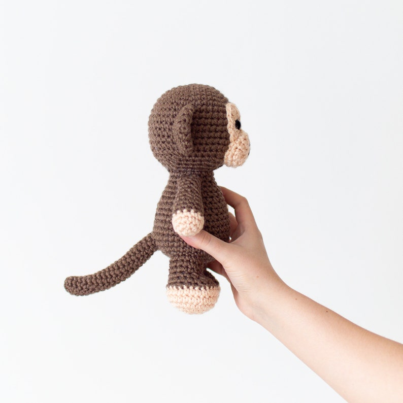 May include: A brown crocheted monkey toy with a long tail. The monkey has a light brown face and a pink nose.
