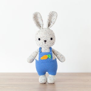 May include: A crocheted bunny toy wearing blue dungarees with a carrot patch. The bunny has white fur with brown speckles and black eyes.