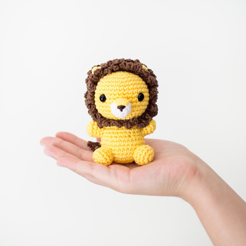 May include: A small crocheted lion with a brown mane, yellow body, and black eyes. The lion is sitting on a hand.
