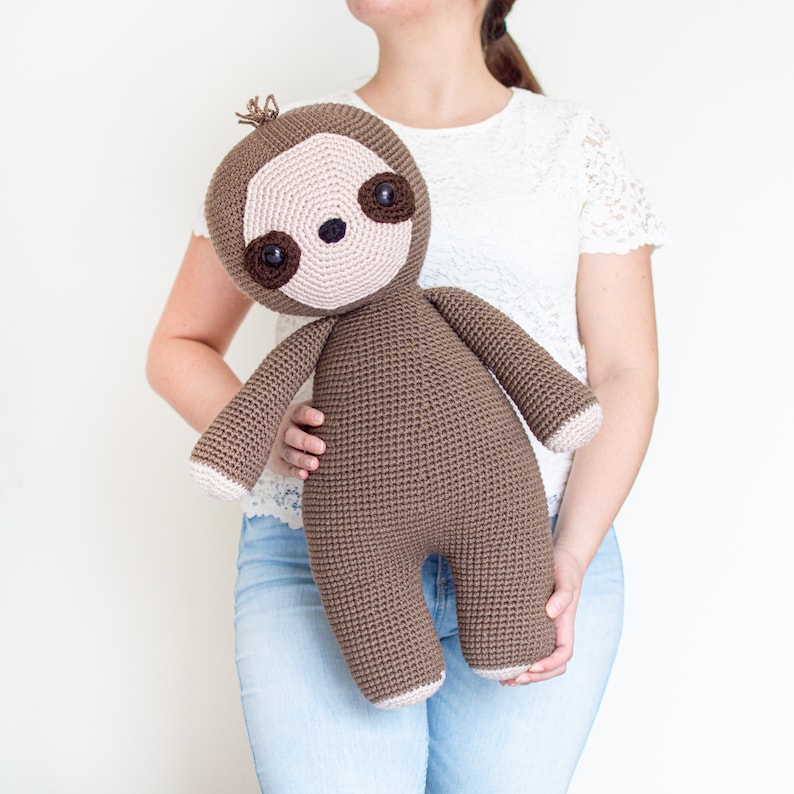 May include: A brown crocheted sloth plush toy with black eyes and a pink nose. The sloth is being held by a person wearing blue jeans and a white lace top.