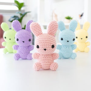May include: Four crocheted bunny rabbits in different pastel colors: light green, purple, pink, and light blue. The bunnies are sitting on a white surface.