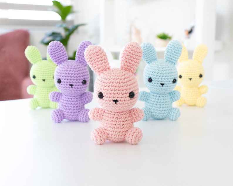 May include: Four crocheted bunny rabbits in different pastel colors: light green, purple, pink, and light blue. The bunnies are sitting on a white surface.