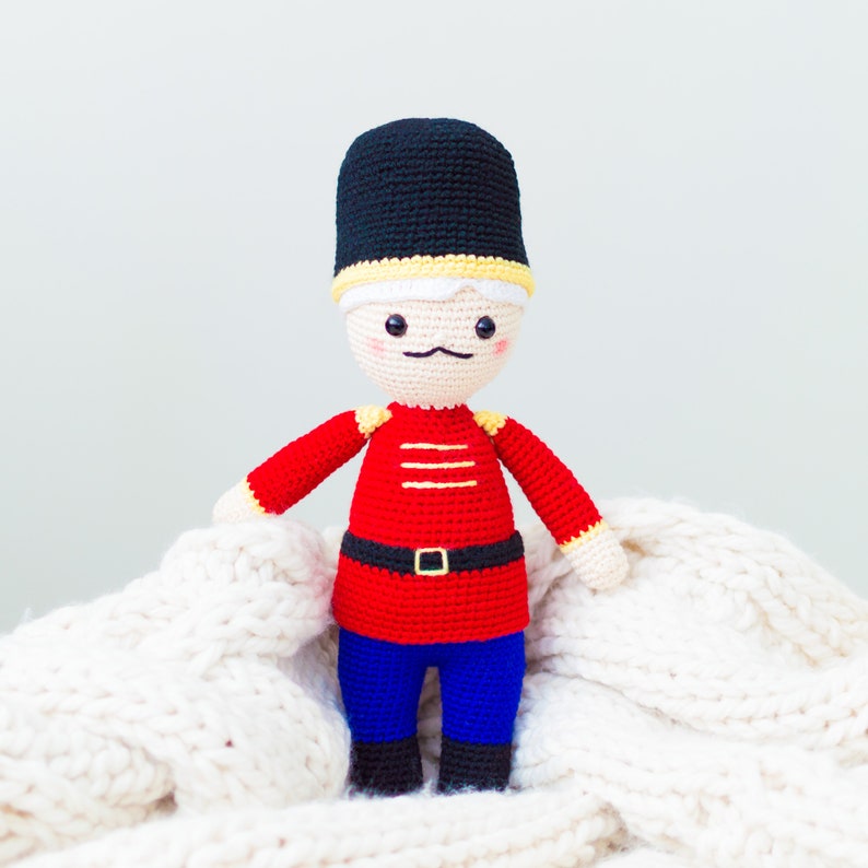 May include: A crocheted toy soldier with a red uniform, yellow buttons, and a black hat. The toy is sitting on a white knitted blanket.