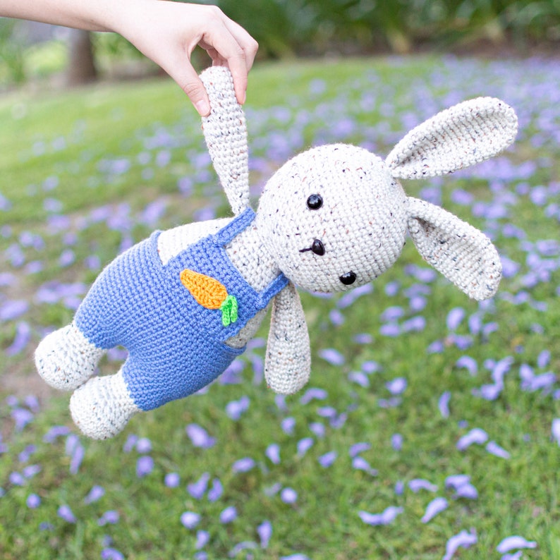 May include: A crocheted bunny toy wearing blue dungarees with a carrot patch. The bunny is white with brown speckles and has black eyes. The bunny is being held by a hand.