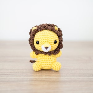 May include: A crocheted lion toy with a brown mane, yellow body, and black eyes. The toy is sitting on a wooden surface.