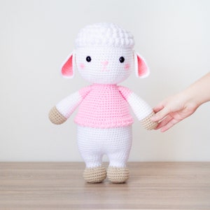 May include: A white crocheted lamb with pink ears and a pink shirt. The lamb has brown crocheted feet and is standing on a wooden surface.