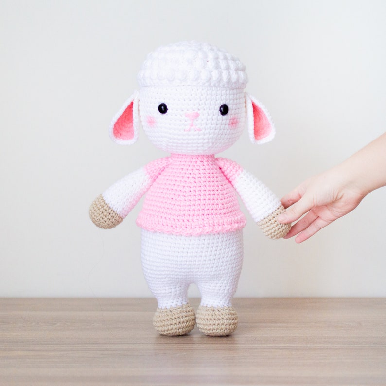 May include: A white crocheted lamb with pink ears and a pink shirt. The lamb has brown crocheted feet and is standing on a wooden surface.