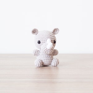 May include: A small gray crocheted rhinoceros toy with black eyes and a white horn. The toy is sitting on a light brown wooden surface.