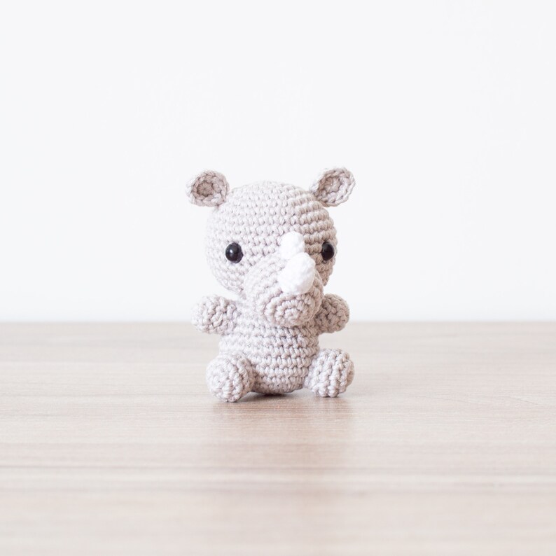 May include: A small gray crocheted rhinoceros toy with black eyes and a white horn. The toy is sitting on a light brown wooden surface.