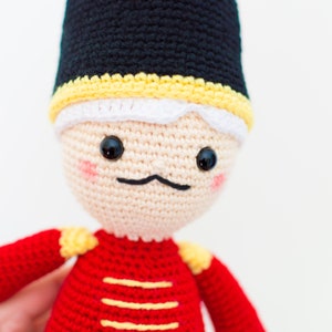 May include: A crocheted toy soldier with a black hat, white trim, and a red and yellow uniform. The toy has a friendly face with black eyes and a black mustache.