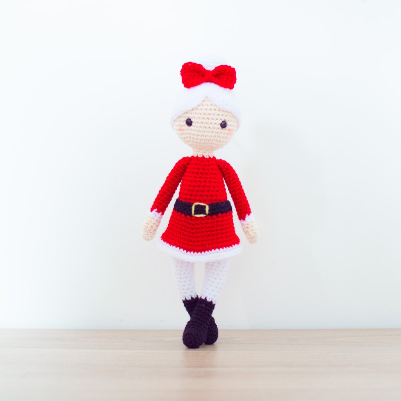 Puede incluir: Una mu&ntilde;eca de crochet vestida de Pap&aacute; Noel. La mu&ntilde;eca lleva un vestido rojo con ribete blanco, un cintur&oacute;n negro y un ribete de piel blanca alrededor del sombrero. La mu&ntilde;eca tiene una cara blanca con ojos negros y un lazo rojo.