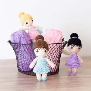 May include: Three crocheted dolls with different colored dresses and hair. The dolls are sitting in a wire basket with balls of yarn.