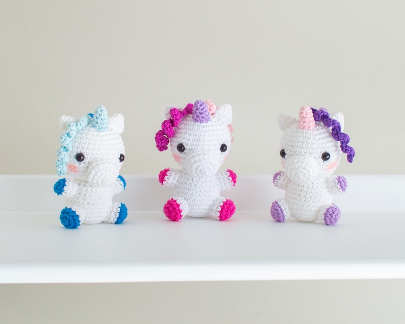 Op de afbeelding: Drie gehaakte eenhoorn speelgoedjes, elk met een andere kleur manen en staart. De eenhoorns zijn wit met roze wangen en zwarte ogen. De kleuren van de manen en staart zijn blauw, roze en paars.