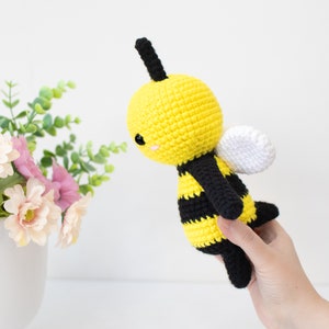 May include: A crocheted bee plush toy with black and yellow stripes, white wings, and black antennae. The bee has a cute, cartoon-like face with black eyes and a pink smile.