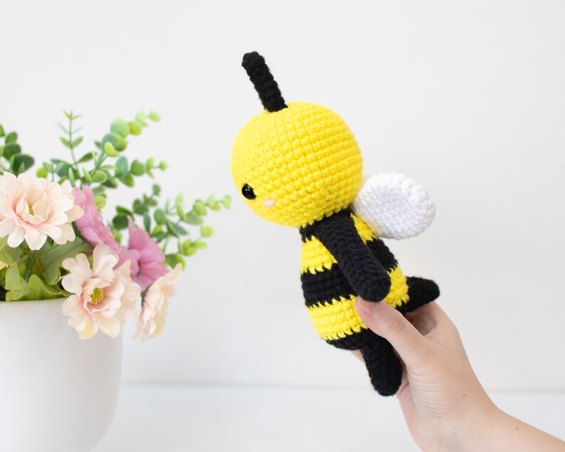 May include: A crocheted bee plush toy with black and yellow stripes, white wings, and black antennae. The bee has a cute, cartoon-like face with black eyes and a pink smile.