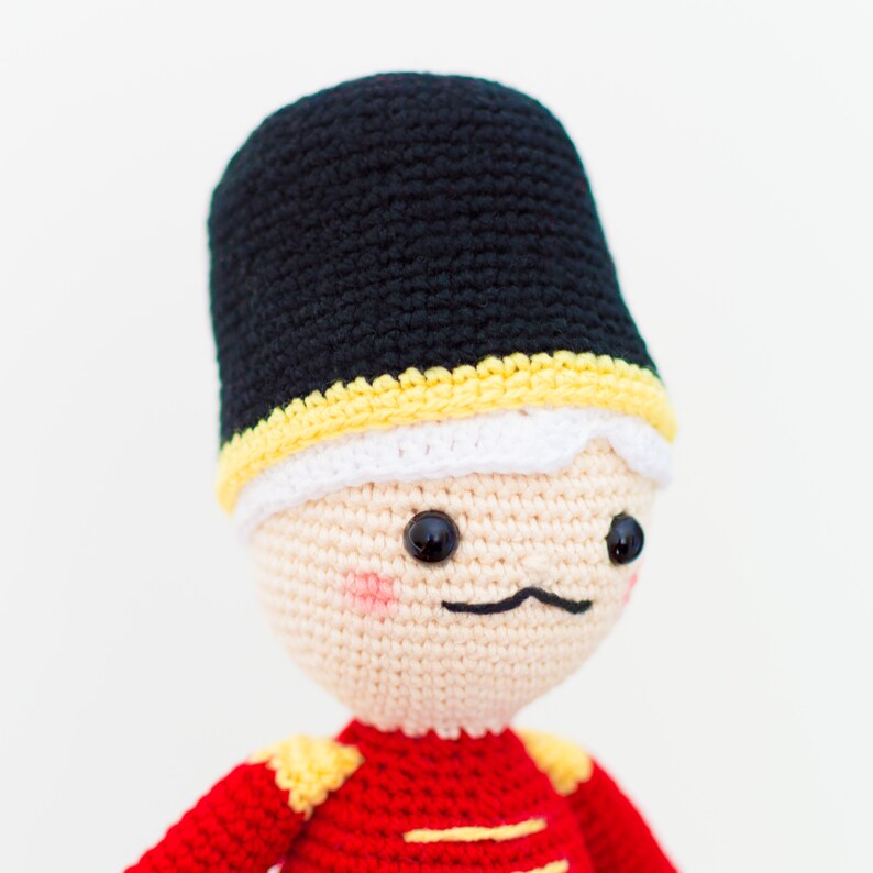 May include: A crocheted toy soldier with a black hat, white trim, and a red and yellow uniform.