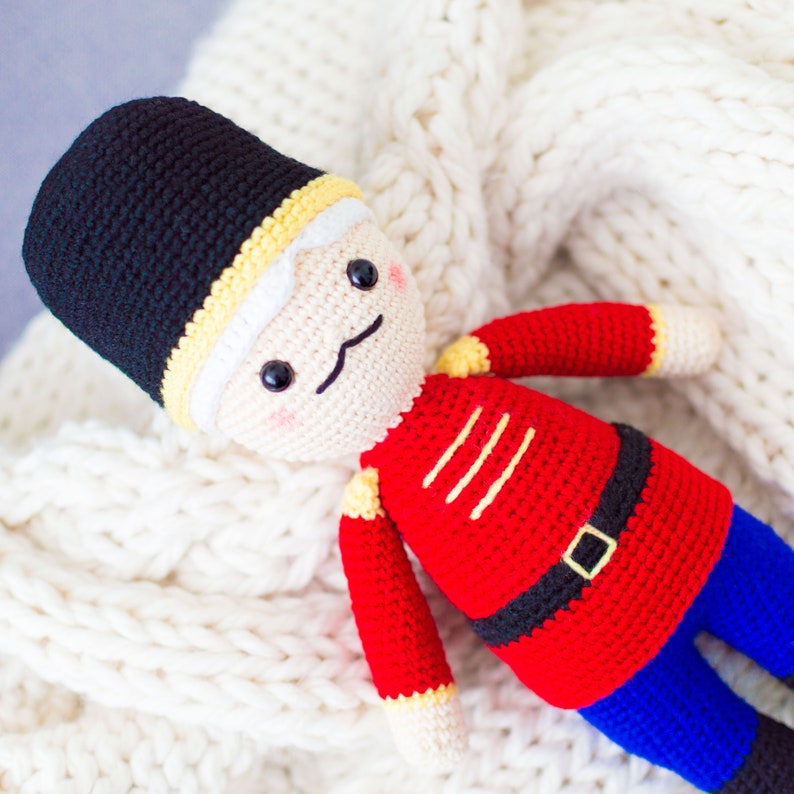 May include: A crocheted toy soldier with a black hat, red uniform, and blue pants. The toy has a white face with painted eyes, a nose, and a mustache.