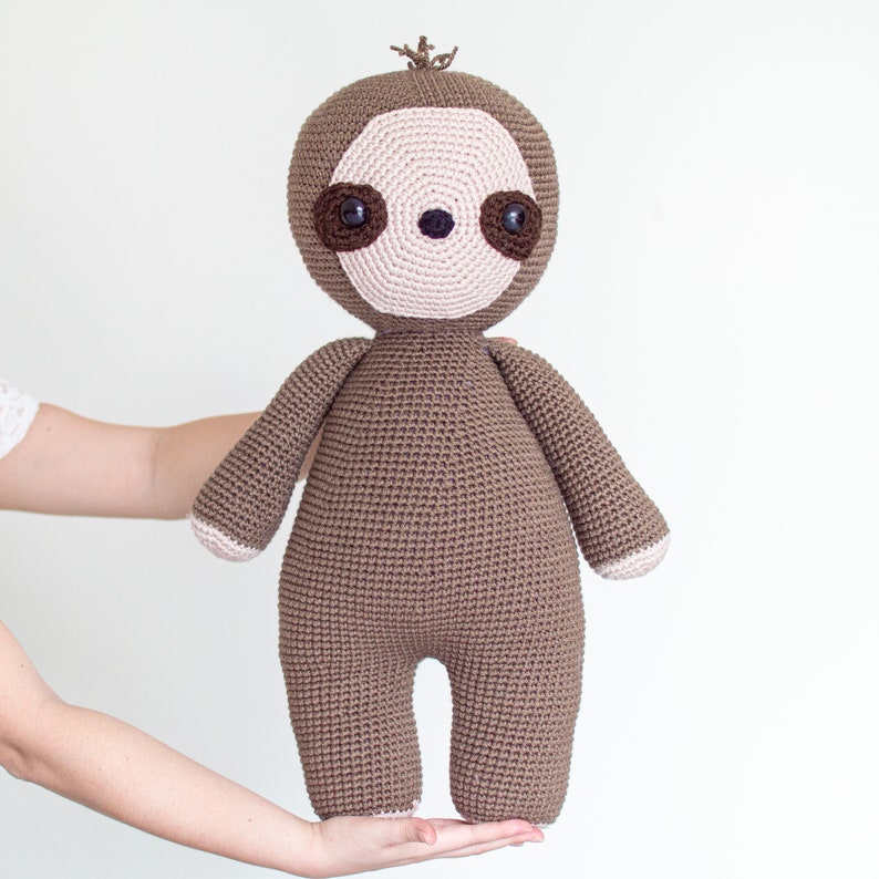 May include: A brown crocheted sloth plush toy with a pink face and brown eyes. The sloth is standing on a person's hand.