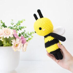 May include: A crocheted bee toy with black and yellow stripes, white wings, and black eyes. The bee is held in a hand against a white background.