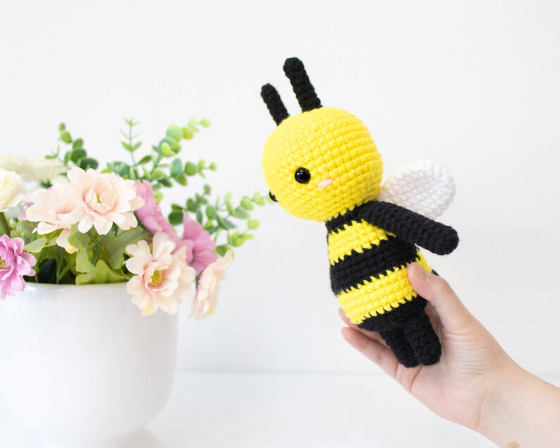 May include: A crocheted bee toy with black and yellow stripes, white wings, and black eyes. The bee is held in a hand against a white background.