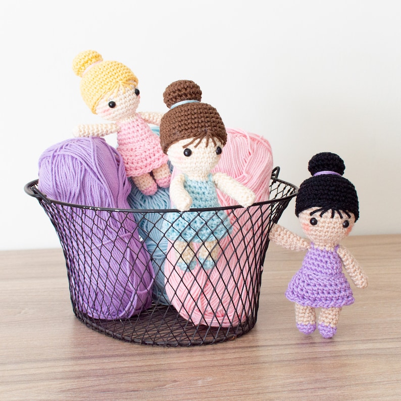 May include: Three crocheted ballerina dolls in a wire basket with pink, purple, and light blue yarn. The dolls are wearing pink, blue, and purple dresses.