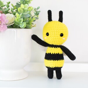 May include: A crocheted bee toy with black and yellow stripes, black legs, and black antennae. The bee has a yellow head with black eyes and a small, white smile.