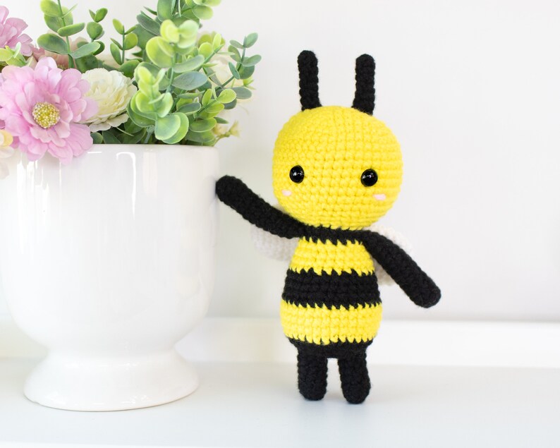 May include: A crocheted bee toy with black and yellow stripes, black legs, and black antennae. The bee has a yellow head with black eyes and a small, white smile.