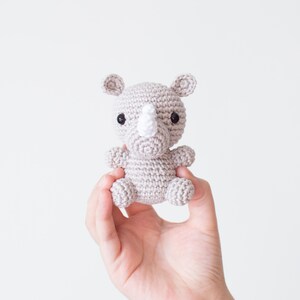 May include: A small gray crocheted rhinoceros toy with a white horn and black eyes. The toy is being held in a person's hand.