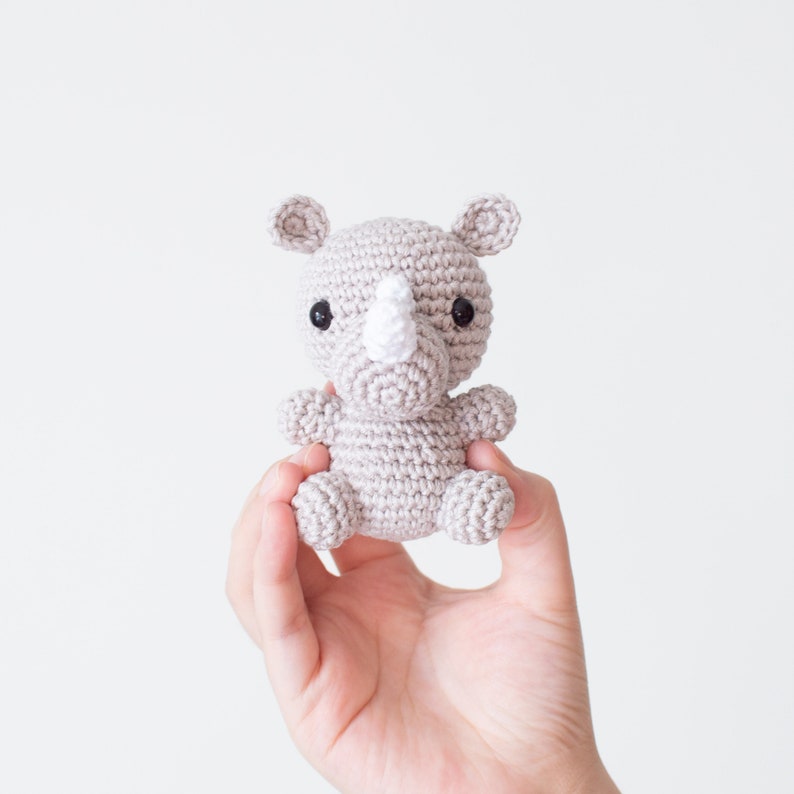 May include: A small gray crocheted rhinoceros toy with a white horn and black eyes. The toy is being held in a person's hand.