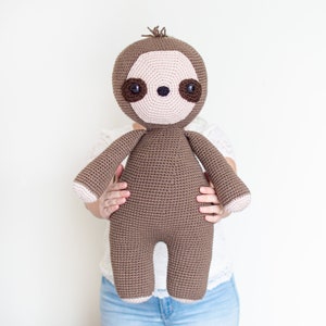 May include: A brown crocheted sloth plush toy with pink facial features and brown eyes. The sloth is being held by a person wearing a white shirt and blue jeans.