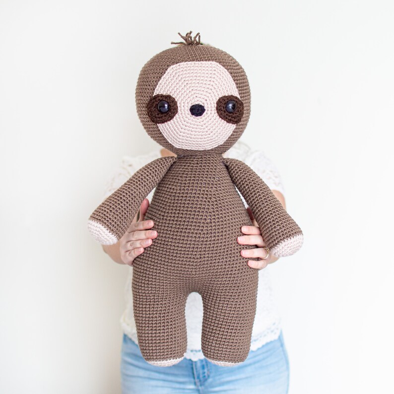 May include: A brown crocheted sloth plush toy with pink facial features and brown eyes. The sloth is being held by a person wearing a white shirt and blue jeans.