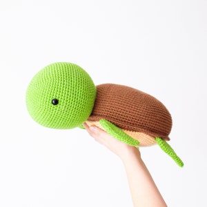 May include: A green and brown crocheted turtle plush toy with a black eye. The turtle is being held in a hand with the head facing the left side of the image.