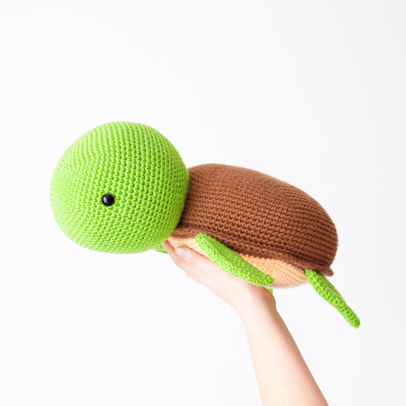 May include: A green and brown crocheted turtle plush toy with a black eye. The turtle is being held in a hand with the head facing the left side of the image.