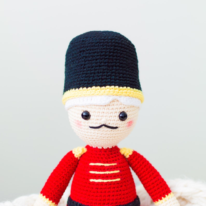 May include: A crocheted toy soldier with a black hat, red and yellow uniform, and a white face with black eyes and a mustache.