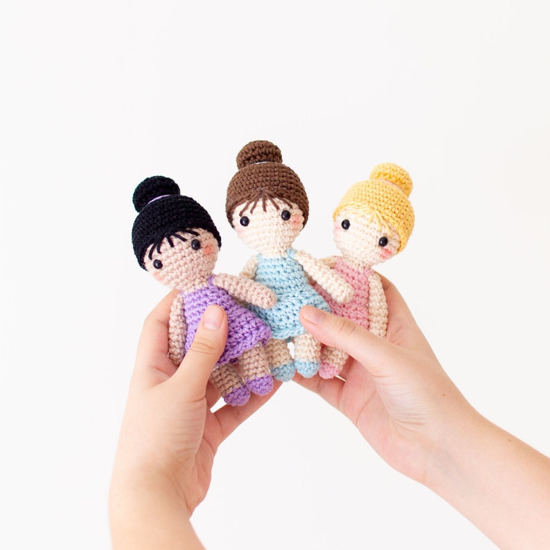 May include: Three crocheted dolls dressed in ballerina outfits. The dolls are wearing pink, blue, and purple dresses. They have brown, black, and blonde hair.