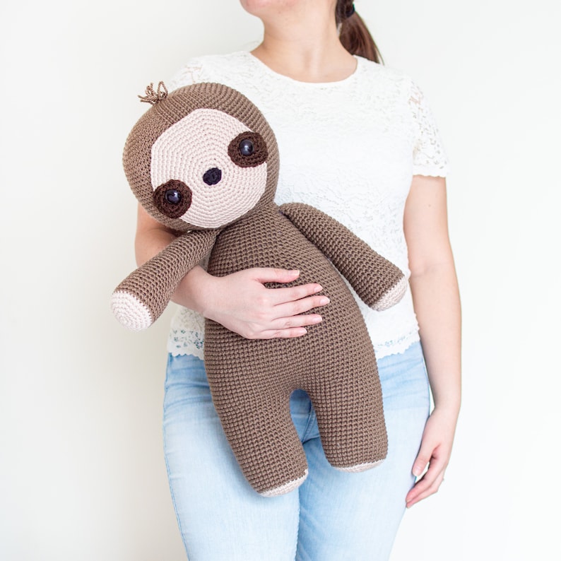 May include: A brown crocheted sloth plush toy with a pink face and brown eyes. The sloth is being held by a person wearing a white lace top and blue jeans.