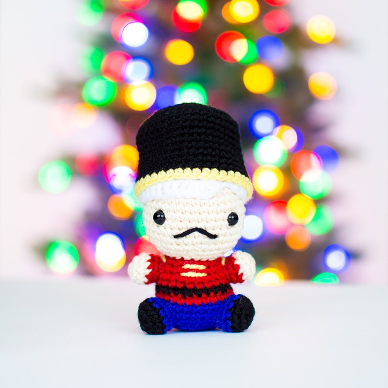 May include: A handmade crochet toy soldier with a black hat, a cream-colored face, and a black mustache. The soldier wears a red and black jacket, blue pants, and black boots. The background is a blurred display of Christmas lights.