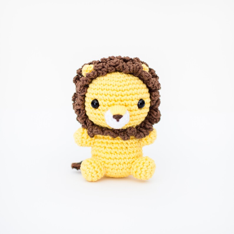 May include: A crocheted lion toy with a yellow body and brown mane. The lion has black eyes and a white nose.