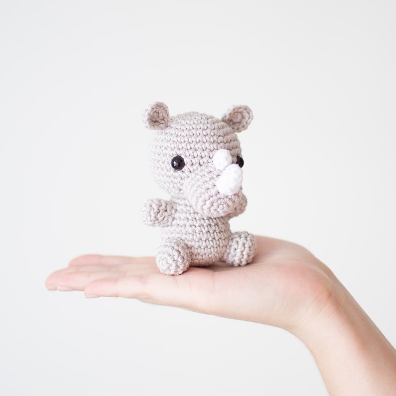 May include: A small, gray crocheted rhinoceros toy with black eyes and a white horn. The toy is sitting in the palm of a hand.