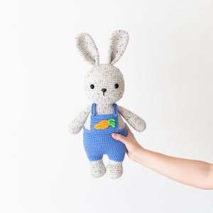 May include: A crocheted white bunny wearing blue dungarees with a green and orange carrot applique. The bunny is being held by a hand.