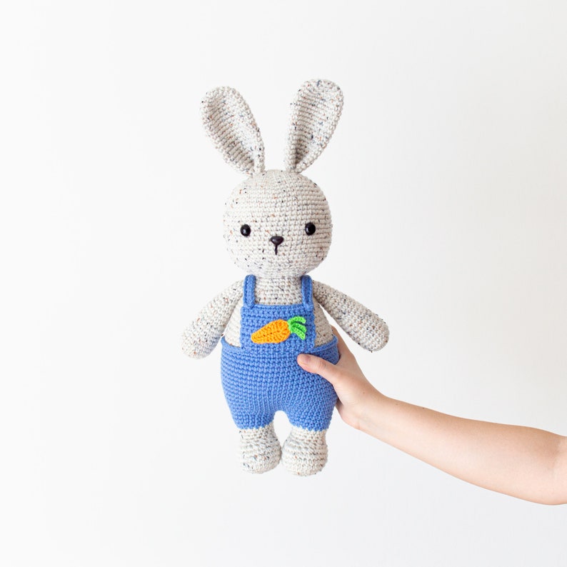 May include: A crocheted white bunny wearing blue dungarees with a green and orange carrot applique. The bunny is being held by a hand.